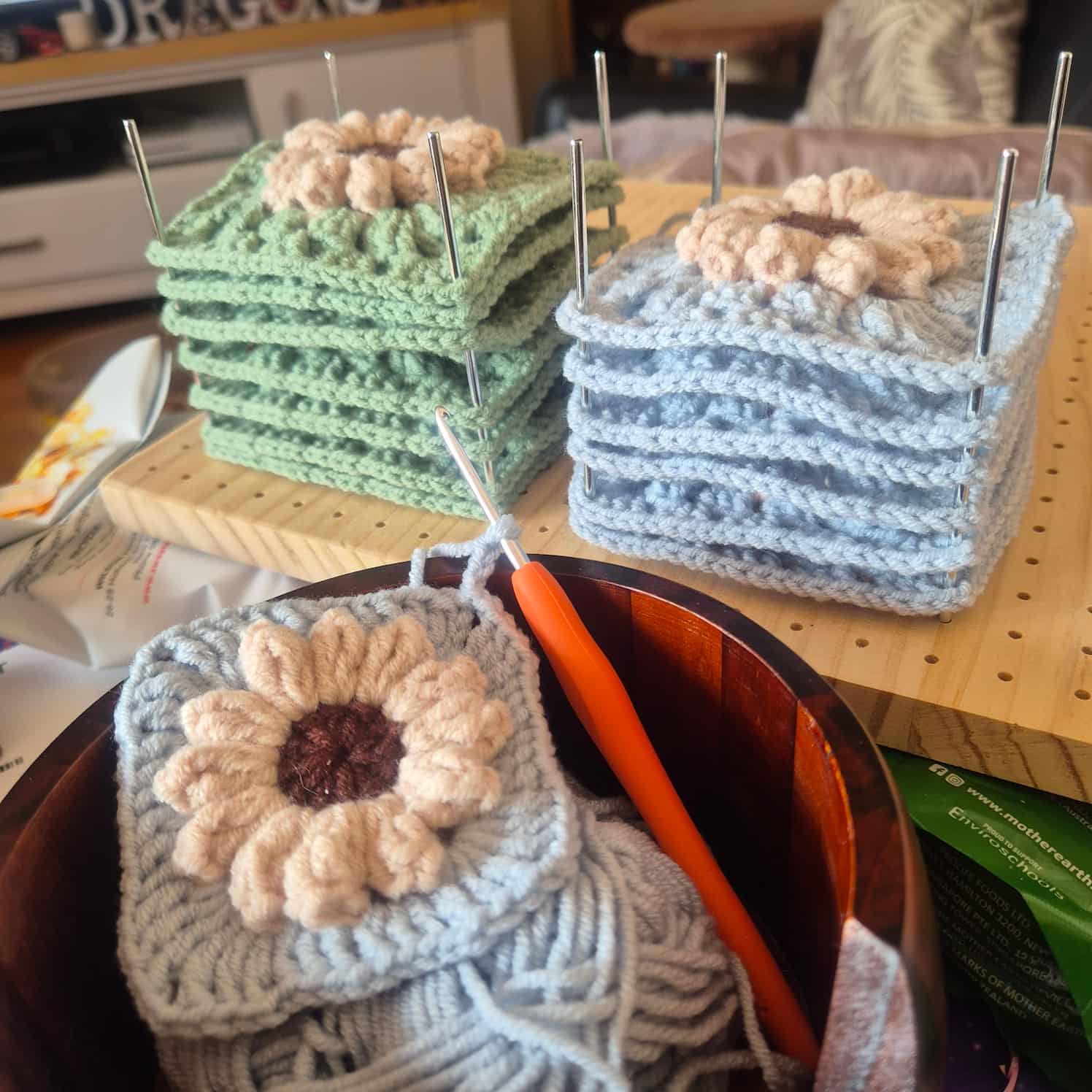 A stack of green and blue granny squares on a pin board. One blue granny square is in the foreground, mostly completed, with a crochet hook still through a stitch. The squares all have a flower with cream petals and brown center.