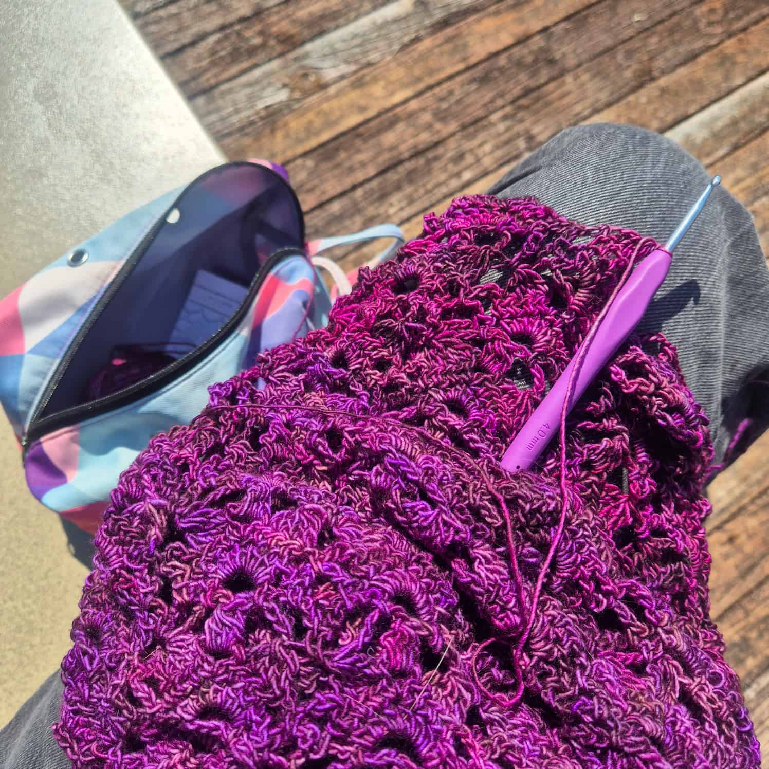 a pinky-purple crochet project is draped over a knee, crochet hook through one stitch. It has an intricate lace pattern.