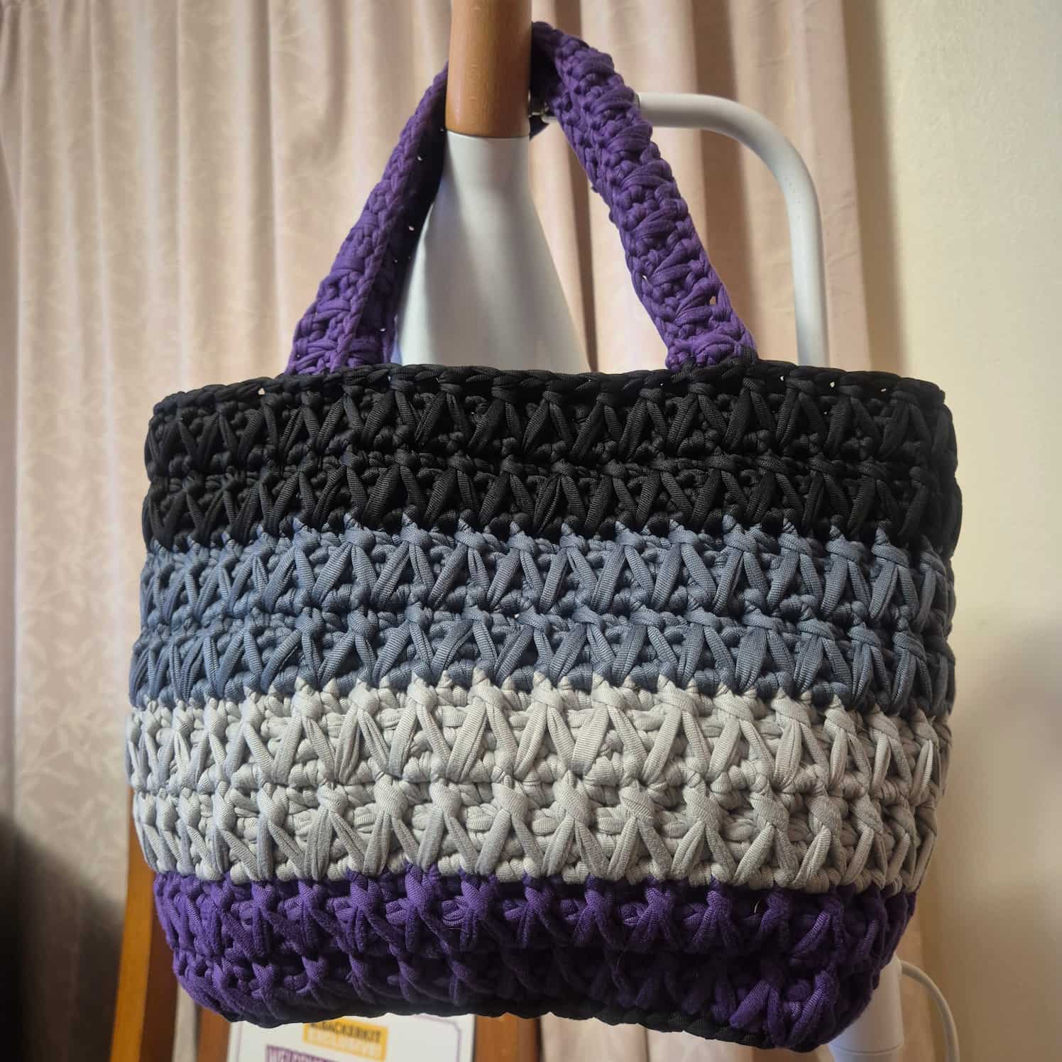 A crochet bag made from t-shirt yarn and in the asexual flag colours - black at the top, then grey, then white (though the bag uses a very light grey), then purple at the bottom. The straps are purple.