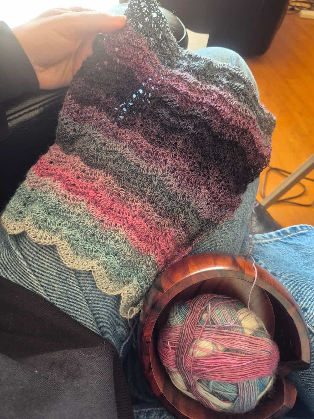 About 30 centimeters of a completed crochet scarf. The pattern is a zig-zag style, and the yarn changes colours from pale to light blue, pink, grey, purple and black.