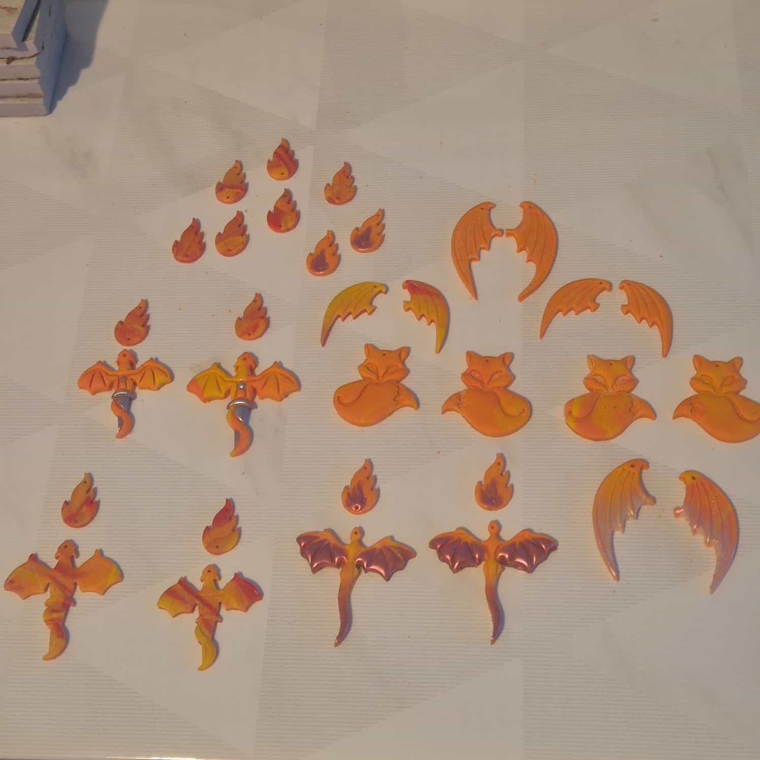 A wide shot of many cut out polymer clay flat designs. Dragons, foxes, wings, and fire. All in orange and yellow.