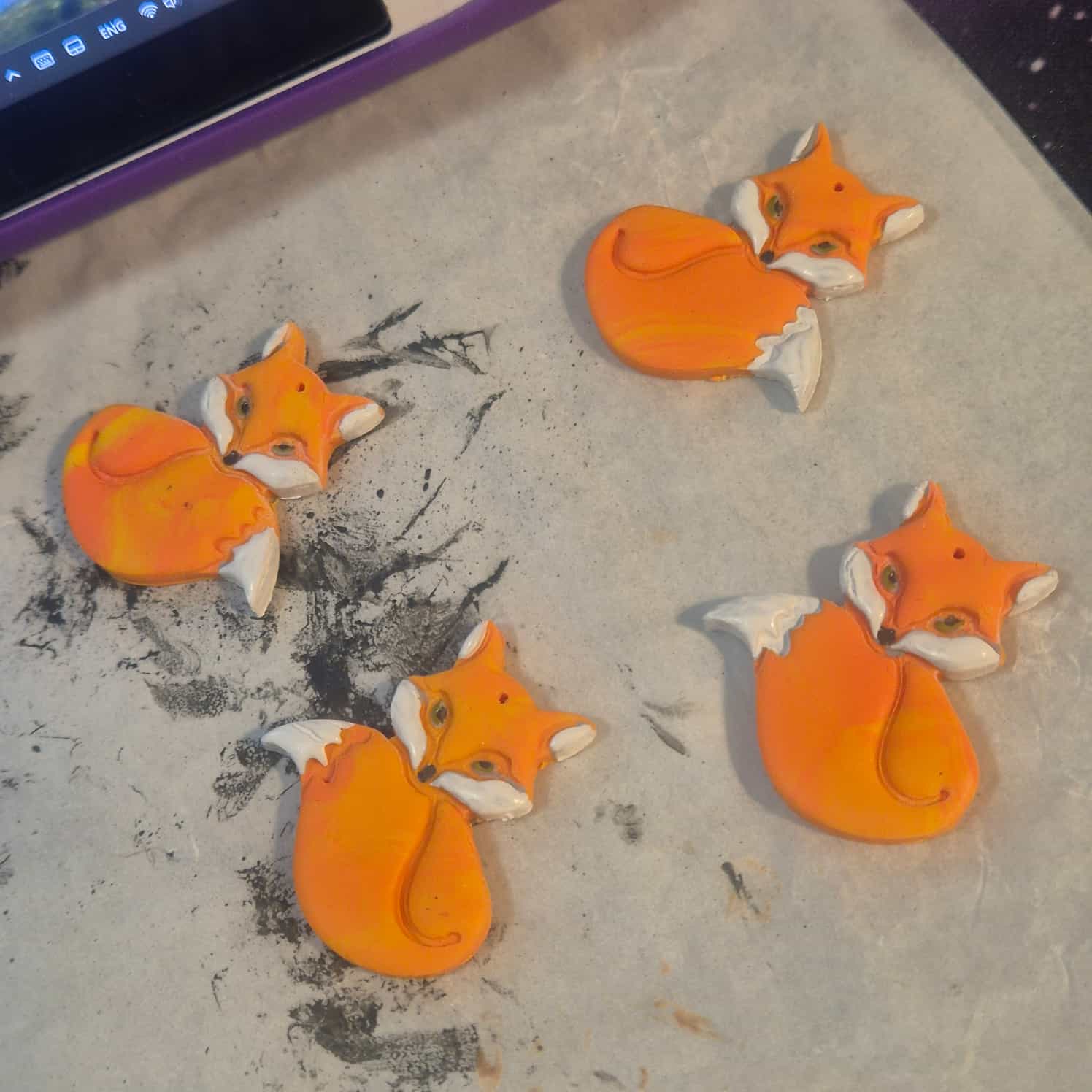 Four orange and yellow polymer clay flat fox designs. Their cheeks, tail tip and ears are painted white.