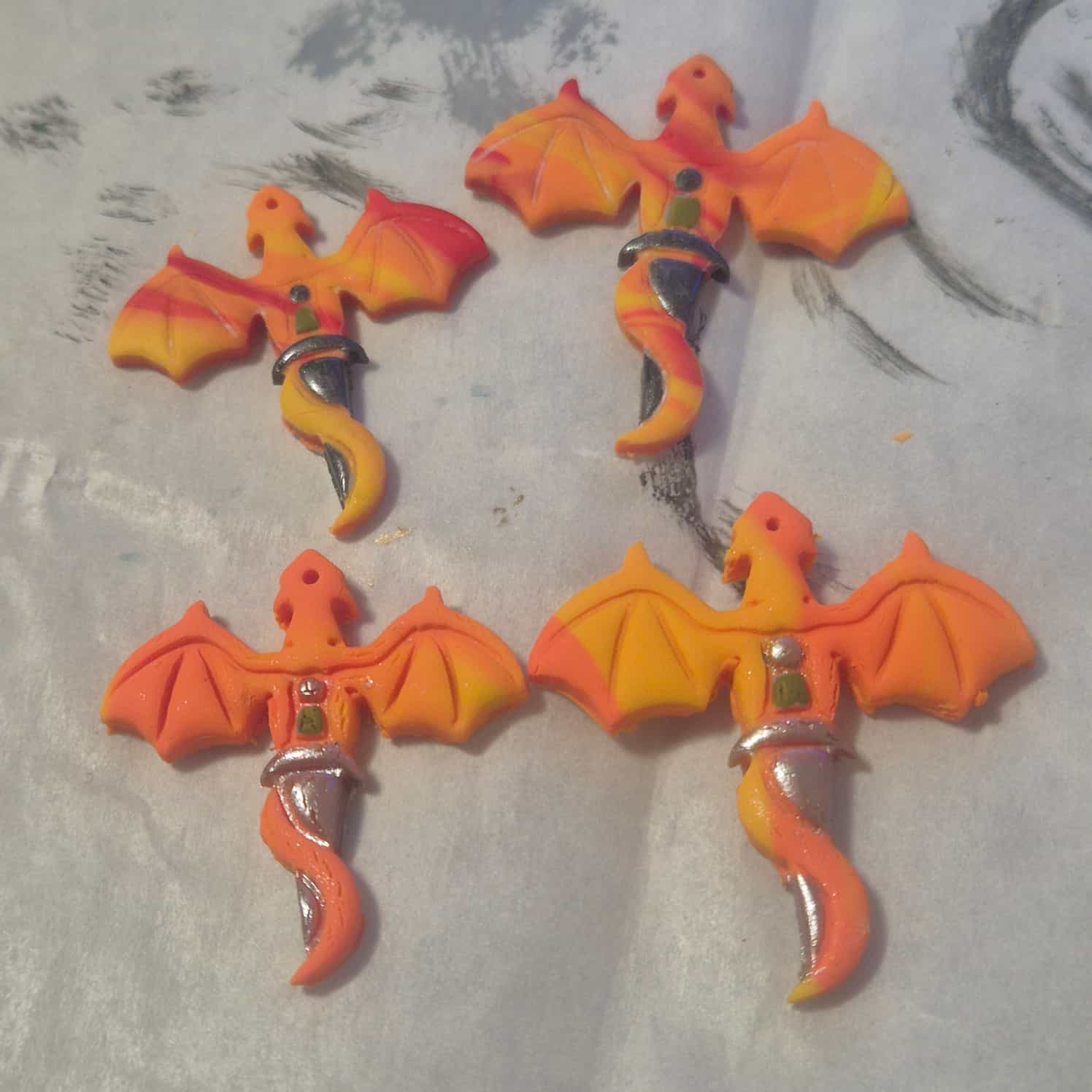 Four orange and yellow polymer clay flat dragon designs. The dragons have their wings spread and their tails wrapped around a sword, which is painted silver.