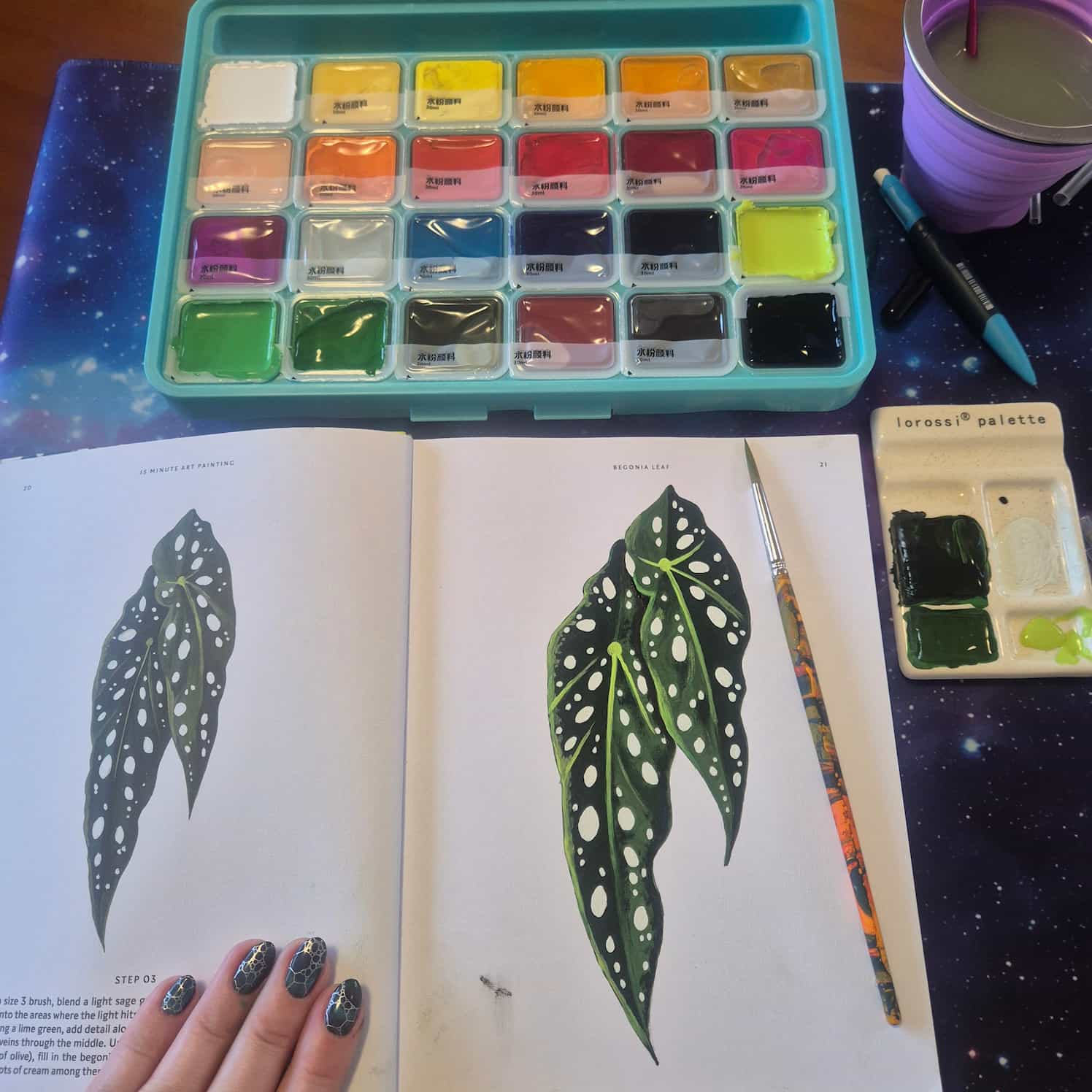A book with the instructions for the painting on the left, and the completed painting on the right. The painting is of begonia leaves, drawn with gouache paints. The paint palette is at the top of the frame, it has 24 colours. The paintbrush sits on the book, and a palette and water cup are off to the right.