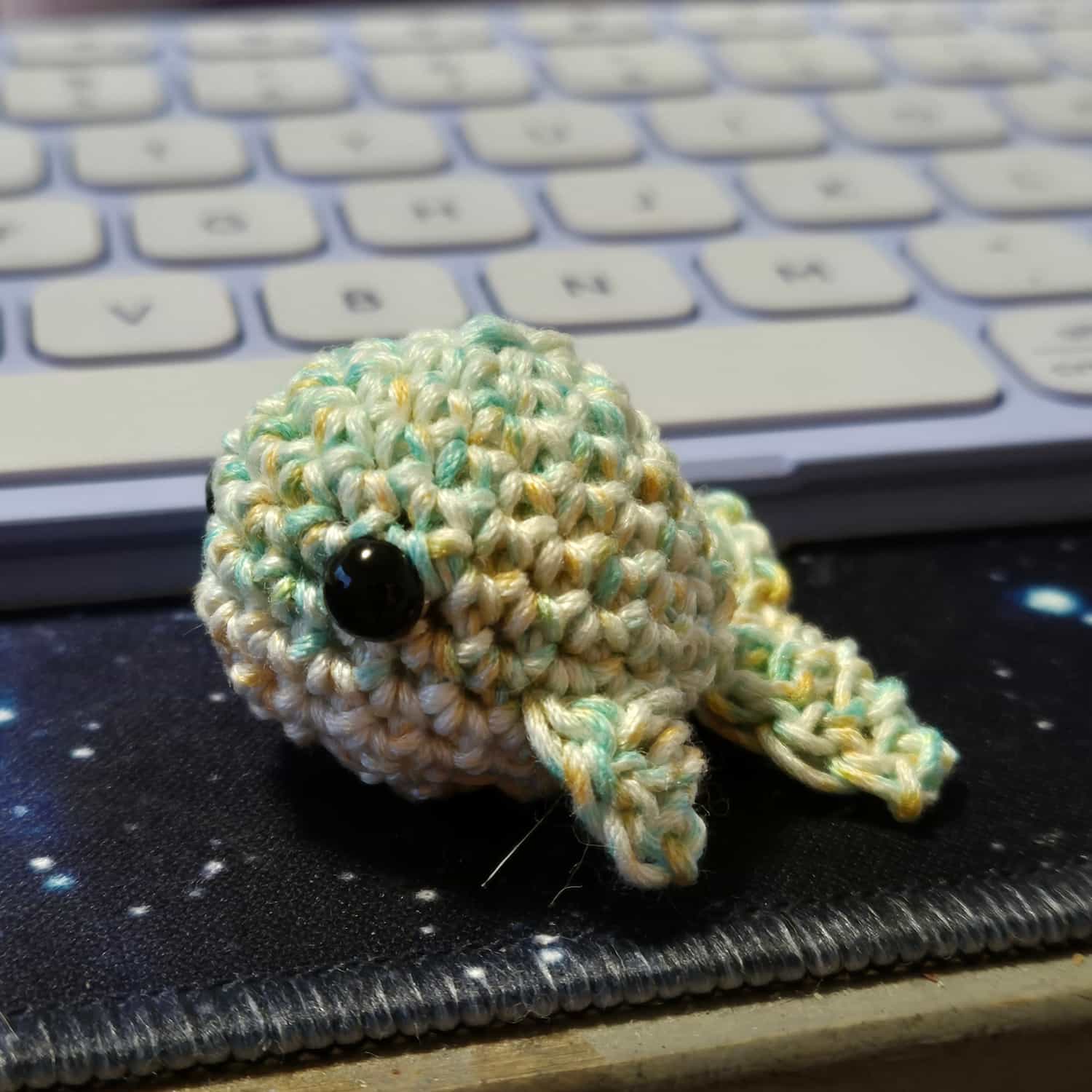 A small crocheted whale. It has a green, yellow and white upper body, small fins and tail. The under body is yellow and white. It has black bead eyes.